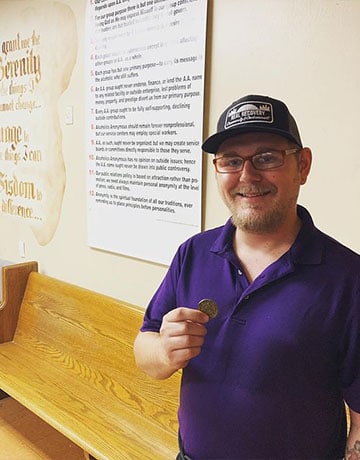 sober living outcomes | Real Recovery alumni holding one year anniversary sobriety coin in front of the 12 steps at an AA meeting